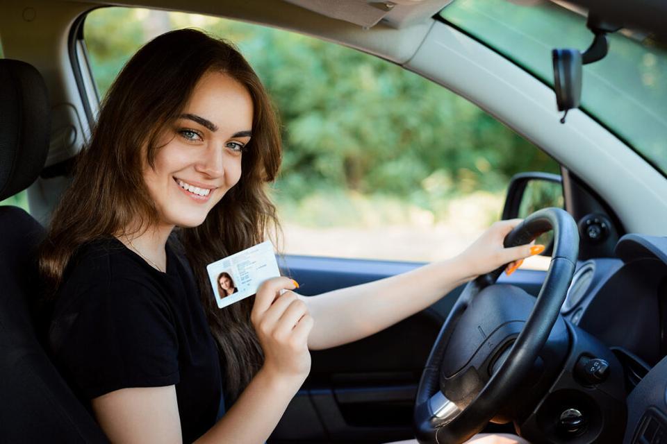 Smiling young female with pleasant appearance shows proudly her drivers license, sits in new car, being young inexperienced driver, looks with joyful expression�