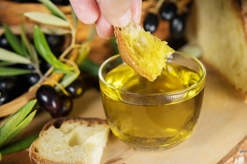 A hand dipping a piece of bread in a bowl full of Extra Virgin Olive Oil made in Puglia, Salento on a wooden table. Tipical ingredients of mediterranean diet, healhty eating concept